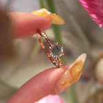 Dainty yellow gold Ilona style one of a kind engagement ring by Laurie Fleming Jewellery held between index finger and thumb and rotated side to side. The ring features a silky seafoam turquoise green sapphire and delicate twinkling diamonds on the band. There are flowers partially visible in the foreground and background, with the ring in focus at the centre.