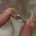 A one of a kind 14k champagne gold "Water Lily" engagement ring by Laurie Fleming Jewellery with a silver-blush colour change Montana sapphire centre held in delicate hand-carved claw prongs. A sprinkling of petite and delicate diamonds dance along the band. The ring is held between an index finger and thumb and rotated gently from side to side. The background, which is blurred and out of focus, is of plants.