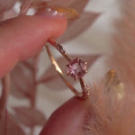 A delicate hand-carved one of a kind "Ilona" style engagement ring by Laurie Fleming Jewellery. The ring features a rose cut pink sapphire centre, a tapered knife-edge band, and small diamond accents on the band shoulders. The ring is held between index finger and thumb of a hand and is rotated from side to side. The background features dried light red leaves and the foreground has a fluffy plant partially visible.