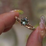 A delicate and ethereal hand-carved one of a kind "Daphne" style engagement ring by Laurie Fleming Jewellery. The ring features a round brilliant cut light green sapphire centre, with clusters of rose and brilliant cut diamond accents on the band in 14k yellow gold. The ring is held between an index finger and thumb and rotated gently from side to side. The background, which is blurred and out of focus, is of plants.