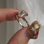 A hand holding a delicate and ethereal hand-carved one of a kind "Daphne" style engagement ring by Laurie Fleming Jewellery between index finger and tumb, rotating it from side to side. The ring features a 1.01ct emerald cut diamond with rose and brilliant cut diamond accents on the band in 14k yellow gold. The background is light grey with some draped silk fabric.