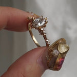 A hand holding a one of a kind "Daphne" style engagement ring by Laurie Fleming jewellery between index finger and thumb, rotating the ring back and forth. The ring features a 0.70ct modified oval cut diamond with a unique glimmering sparkle, with diamond clusters on the band. The background is a light gray with some draped silk fabric.