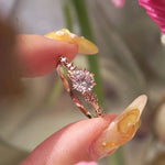 A one of a kind "Daphne" style engagement ring by Laurie Fleming Jewellery,held between index finger and thumb and rotated side to side.  The ring features a pale rosewater pink sapphire centre. The band is encrusted in sparkling diamond clusters. The ring is handcarved and made in solid champagne yellow gold. There are partially visible flowers in the foreground and the background, with the focus on the ring at the centre of the frame.