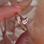 A delicate and ethereal hand-carved one of a kind "Daphne" style engagement ring by Laurie Fleming Jewellery. The ring features an emerald cut peach-pink Montana sapphire centre, with clusters of rose and brilliant cut diamond accents on the band in solid 14k yellow gold. The ring is held between index finger and thumb of a hand and is rotated from side to side. The background features dried light red leaves and the foreground has a fluffy plant partially visible.