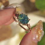 A delicate and ethereal "Nereid" one of a kind engagement ring by Laurie Fleming Jewellery, with a unique opalescent silky teal/turquoise/blue sapphire in an oval cut. The band is adorned with rose and brilliant cut diamonds and the ring is made in solid 14k yellow gold. The ring is held between index finger and thumb and rotated slowly from side to side. In the background, there are out-of-focus plants rustling in the breeze.