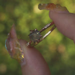 A dainty yellow gold Ilona style engagement ring with a rose cut green sapphire and diamonds on the band, held between index finger and thumb and rotated gently over a background of soft unfocused greenery.