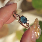 A delicate and ethereal "Nereid" one of a kind engagement ring by Laurie Fleming Jewellery, with a unique opalescent silky teal/turquoise/blue sapphire in a geometric oval cut. The band is adorned with rose and brilliant cut diamonds and the ring is made in solid 14k champagne gold. The ring is held between index finger and thumb and rotated slowly from side to side. In the background, there are out-of-focus plants rustling in the breeze.