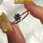 A delicate "Ilona" style one of a kind engagement ring by Laurie Fleming Jewellery in solid 14k champagne gold. It features a stunning sky blue colored round sapphire, held by six hand-shaped claw prongs. The band features small diamonds on the shoulders. The ring is held between an index finger and thumb, and rotated gently from side to side. The background is a pale gray with flecks of rainbow light.