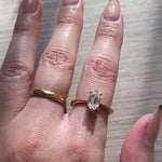 A hand wearing a one of a kind "Aurelia" style ring by Laurie Fleming Jewellery, featuring a unique elongated oval diamond with antique-inspired facets, on a gently tapered and rounded solid gold band. The hand shifts to show the way the diamond's facets sparkle. The background is a light-grey stained wood.