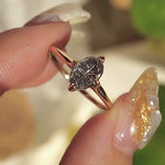 A completely one of a kind, handcarved "Moondew" engagement ring by Laurie Fleming Jewellery. The ring features an oval Starbrite-cut colour change sapphire by lapidary artist John Dyer. The stone is held in four delicate claws, resting atop a gently tapered shining gold band. The ring is held between index finger and thumb and rotated slowly from side to side. In the background, there are out-of-focus plants rustling in the breeze.
