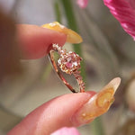 A delicate, one of a kind "Nereid" style engagement ring by Laurie Fleming Jewellery in yellow gold. The ring features a round rose cut light pink sapphire held by four dainty claws, nestled in hand-carved floral peony petals. Rose cut and brilliant diamonds drip down the band. The ring is held between index finger and thumb and rotated from side to side. In the foreground and background flowers are partially visible fluttering in the wind.