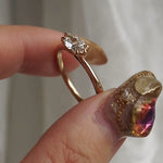 A hand holding a one of a kind "Aurelia" style engagement ring by Laurie Fleming Jewellery between index finger and thumb, rotating it side to side. The ring features a unique "moval" (marquise/oval hybrid) cut diamond set East-West, on a gently tapered and rounded solid gold band. The background is a light grey with some draped silk fabric.