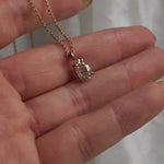 A hand holding a one of a kind, handcarved solid 14k yellow gold necklace featuring a 1.07ct antique pear cut champagne diamond. The diamond is held in four delicate claw-shaped prongs. The pendant hangs from an adjustable 16-18" chain, and is draped across the fingers, which move it gently to show off the diamond's sparkle. In the background, some white gossamer silk fabric hangs.