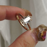 A hand holding a one of a kind "Aurelia" style ring by Laurie Fleming Jewellery between index finger and thumb, rotating it from side to side. The ring features a unique elongated oval diamond with antique-inspired facets, on a gently tapered and rounded solid gold band. The background is light grey with some draped silk fabric.