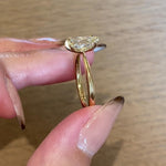 A hand holding a one of a kind "Aurelia" style ring by Laurie Fleming Jewellery between index finger and thumb, showing the side view of the ring and turning it slightly back and forth. The ring features a unique elongated oval diamond with antique-inspired facets, on a gently tapered and rounded solid gold band. The hand shifts to show the way the diamond's facets sparkle. The background is a light-grey stained wood.