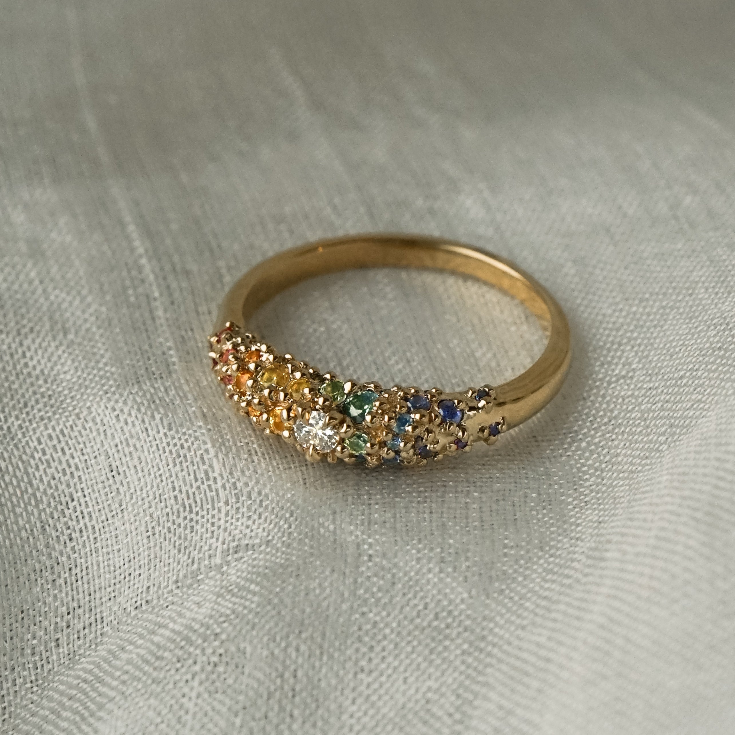 Laurie Fleming Jewellery's "Rainbow Sapphire Moss Signet Ring," a bold solid gold signet-style ring encrusted with sapphires in varying shades and sizes in deep pink, medium pink, light pink, orange, yellow, green, blue-green, medium blue, light blue, and purple, with a single round brilliant cut diamond at the centre on the band. The ring is resting on white gossamer silk.