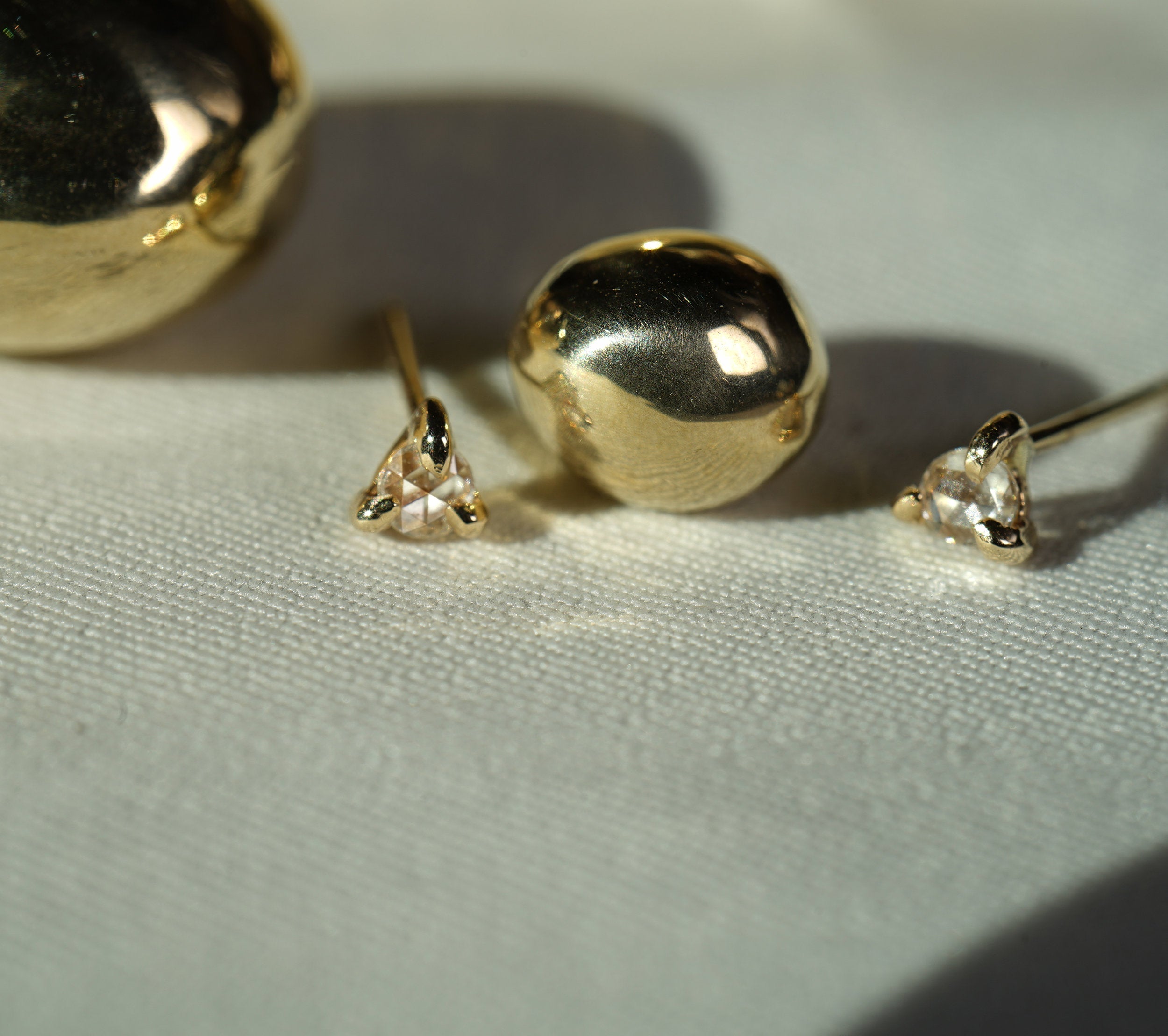 A close up of a pair of Selene stud earrings by Laurie Fleming Jewellery. They each feature a single rose cut diamond held by three claw shaped prongs. They are positioned with decorative balls of gold against white fabric.