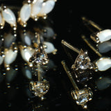 A black mirrored surface with a scattering of stud earrings on it. Towards the back is a single Lyrie earring with marquise-cut mother of pearl. On the middle right, a single Nereid stud with a marquise cut mother of pearl. In the centre, two Selene studs with rose cut diamonds held in three claw-shaped prongs. All earrings are handmade by Laurie Fleming Jewellery.