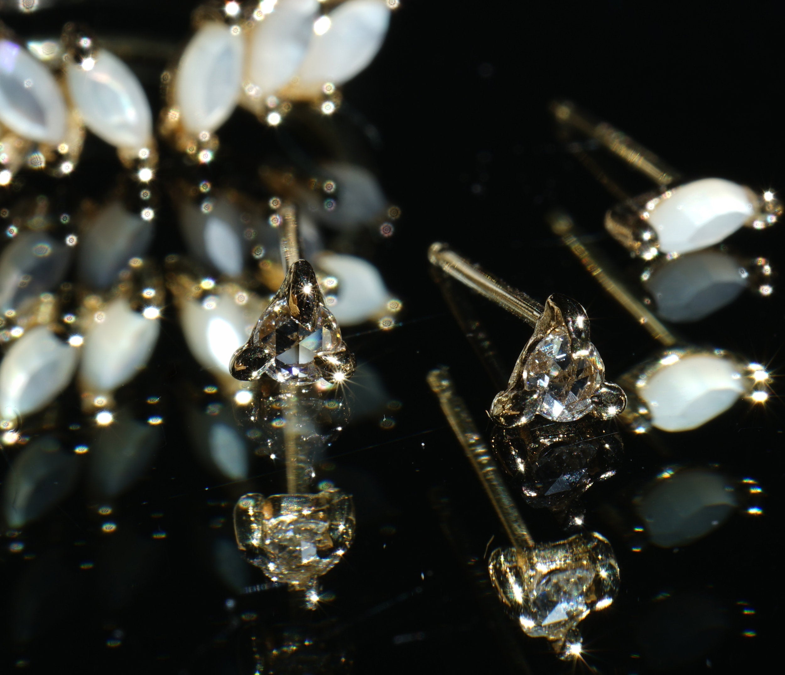 A black mirrored surface with a scattering of stud earrings on it. Towards the back is a single Lyrie earring with marquise-cut mother of pearl. On the middle right, a single Nereid stud with a marquise cut mother of pearl. In the centre, two Selene studs with rose cut diamonds held in three claw-shaped prongs. All earrings are handmade by Laurie Fleming Jewellery.