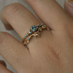 A close up of a hand wearing two rings stacked together. The top ring is a Sapphire Cyndra Ring, Supreme by Laurie Fleming Jewellery featuring teal sapphires along the band and delicate claw-shaped prongs. The bottom ring is a Sapphire Evening Fleur ring, which is a sculptural carved floral band with teal sapphire accents. Both rings are solid gold. The background is light grey.