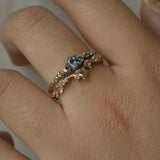 A close up of a hand wearing two rings stacked together. The top ring is a Sapphire Cyndra Ring, Supreme by Laurie Fleming Jewellery featuring teal sapphires along the band and delicate claw-shaped prongs. The bottom ring is a Sapphire Evening Fleur ring, which is a sculptural carved floral band with teal sapphire accents. Both rings are solid gold. The background is light grey.
