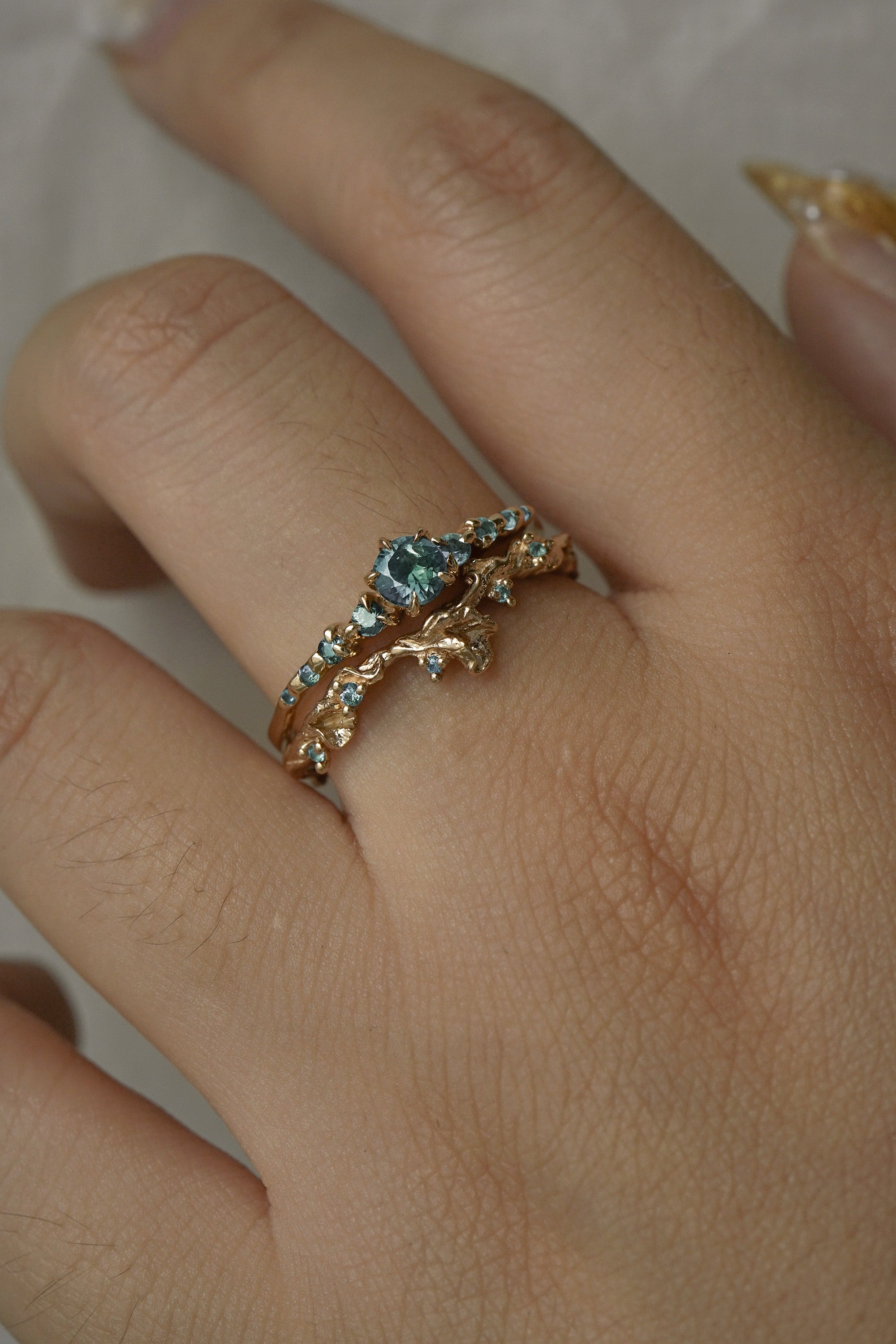 A close up of a hand wearing two rings stacked together. The top ring is a Sapphire Cyndra Ring, Supreme by Laurie Fleming Jewellery featuring teal sapphires along the band and delicate claw-shaped prongs. The bottom ring is a Sapphire Evening Fleur ring, which is a sculptural carved floral band with teal sapphire accents. Both rings are solid gold. The background is light grey.