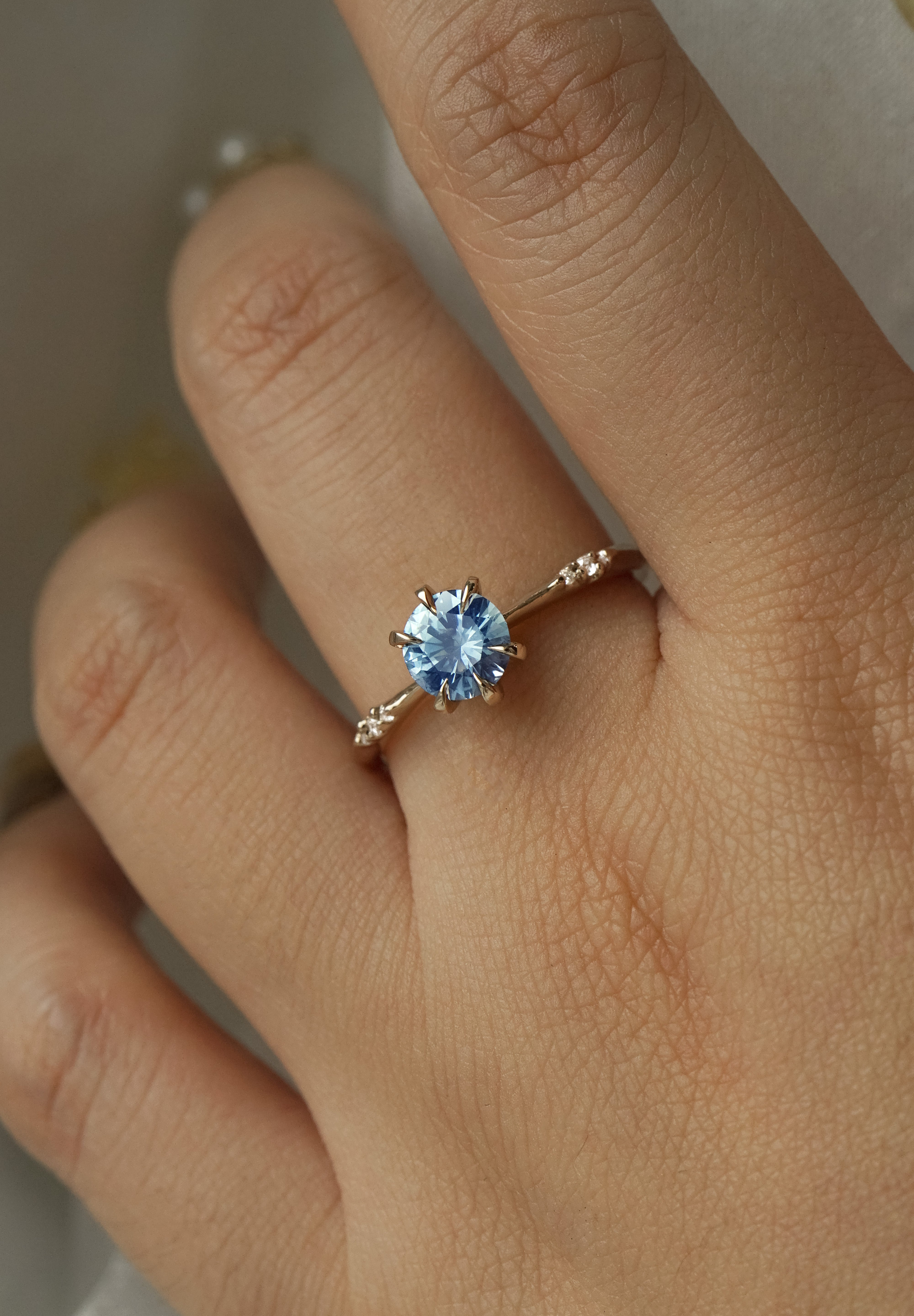 A hand wearing a delicate "Ilona" style one of a kind engagement ring by Laurie Fleming Jewellery in solid 14k champagne gold. It features a stunning sky blue colored round sapphire, held by six hand-shaped claw prongs. The band features small diamonds on the shoulders. The background is a light grey.