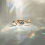 Three one a kind engagement rings by Laurie Fleming Jewellery on a light gray background. On the left, an Ilona style that features an elongated oval shaped scissor cut diamond on a delicate band with diamonds on the shoulders. In the middle, another Ilona ring, this one with a sky blue round sapphire centre stone. On the right, a custom carved Moondew ring featuring a unique long oval diamond. Beams of rainbow light surround the ring.