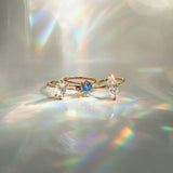 Three one a kind engagement rings by Laurie Fleming Jewellery on a light gray background. On the left, an Ilona style that features an elongated oval shaped scissor cut diamond on a delicate band with diamonds on the shoulders. In the middle, another Ilona ring, this one with a sky blue round sapphire centre stone. On the right, a custom carved Moondew ring featuring a unique long oval diamond. Beams of rainbow light surround the ring.