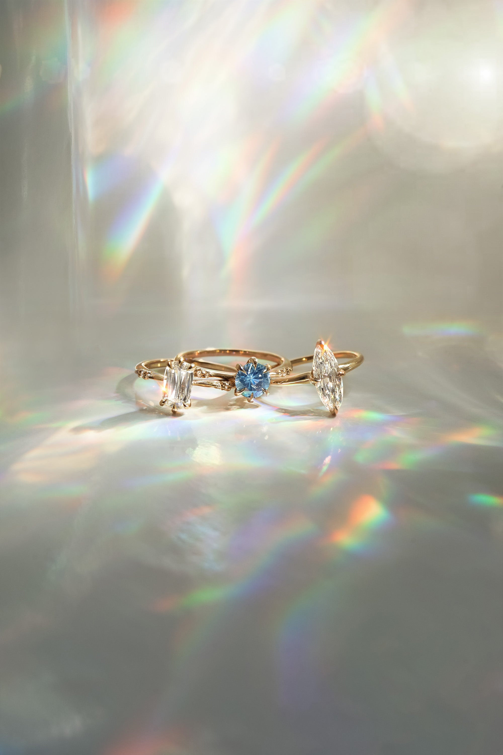 Three one a kind engagement rings by Laurie Fleming Jewellery on a light gray background. On the left, an Ilona style that features an elongated oval shaped scissor cut diamond on a delicate band with diamonds on the shoulders. In the middle, another Ilona ring, this one with a sky blue round sapphire centre stone. On the right, a custom carved Moondew ring featuring a unique long oval diamond. Beams of rainbow light surround the ring.