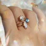 A hand wearing three one of a kind engagement rings by Laurie Fleming Jewellery. The top ring is an Ilona style and features an elongated oval shaped scissor cut diamond on a delicate band with diamonds on the shoulders. In the middle, another Ilona ring, this one with a sky blue round sapphire centre stone. At the base of the finger, a custom carved Moondew ring featuring a unique long oval diamond. The background is light grey with rainbow flecks, and the rings are framed by rainbow curved lens flares.