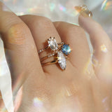 A hand wearing three one of a kind engagement rings by Laurie Fleming Jewellery. The top ring is an Ilona style and features an elongated oval shaped scissor cut diamond on a delicate band with diamonds on the shoulders. In the middle, another Ilona ring, this one with a sky blue round sapphire centre stone. At the base of the finger, a custom carved Moondew ring featuring a unique long oval diamond. The background is light grey with rainbow flecks, and the rings are framed by rainbow curved lens flares.