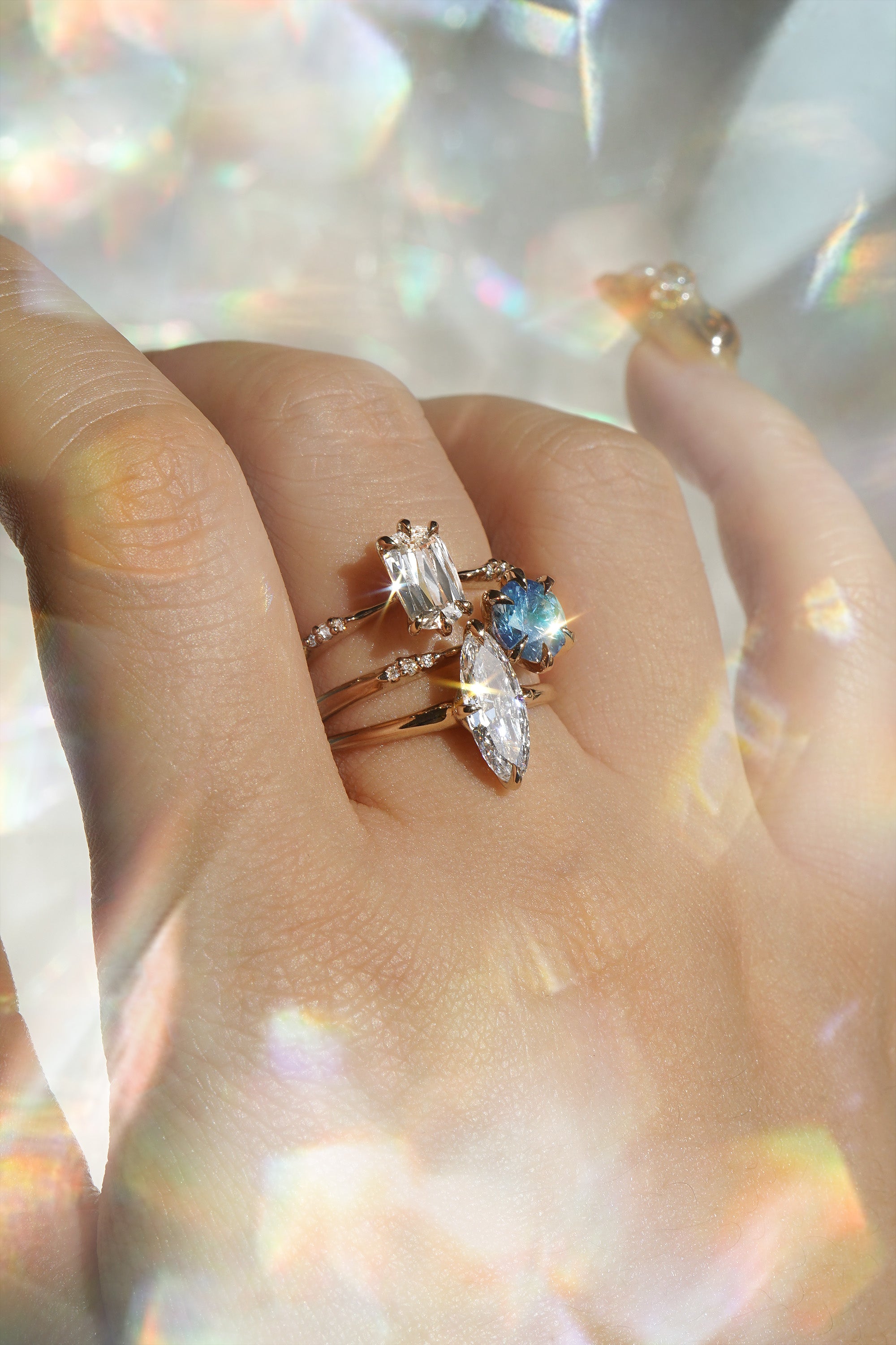 A hand wearing three one of a kind engagement rings by Laurie Fleming Jewellery. The top ring is an Ilona style and features an elongated oval shaped scissor cut diamond on a delicate band with diamonds on the shoulders. In the middle, another Ilona ring, this one with a sky blue round sapphire centre stone. At the base of the finger, a custom carved Moondew ring featuring a unique long oval diamond. The background is light grey with rainbow flecks, and the rings are framed by rainbow curved lens flares.