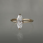 A one of a kind handcarved "Moondew" style engagement ring by Laurie Fleming Jewellery in solid 14k champagne gold. The ring features a unique moval shaped diamond (a long, marquise-oval mix) on a thoughtfully tapered plain gold band, held securely by four prongs at North, South, East, and West. The ring is situated on a medium grey background.
