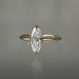A one of a kind handcarved "Moondew" style engagement ring by Laurie Fleming Jewellery in solid 14k champagne gold. The ring features a unique moval shaped diamond (a long, marquise-oval mix) on a thoughtfully tapered plain gold band, held securely by four prongs at North, South, East, and West. The ring is situated on a medium grey background.