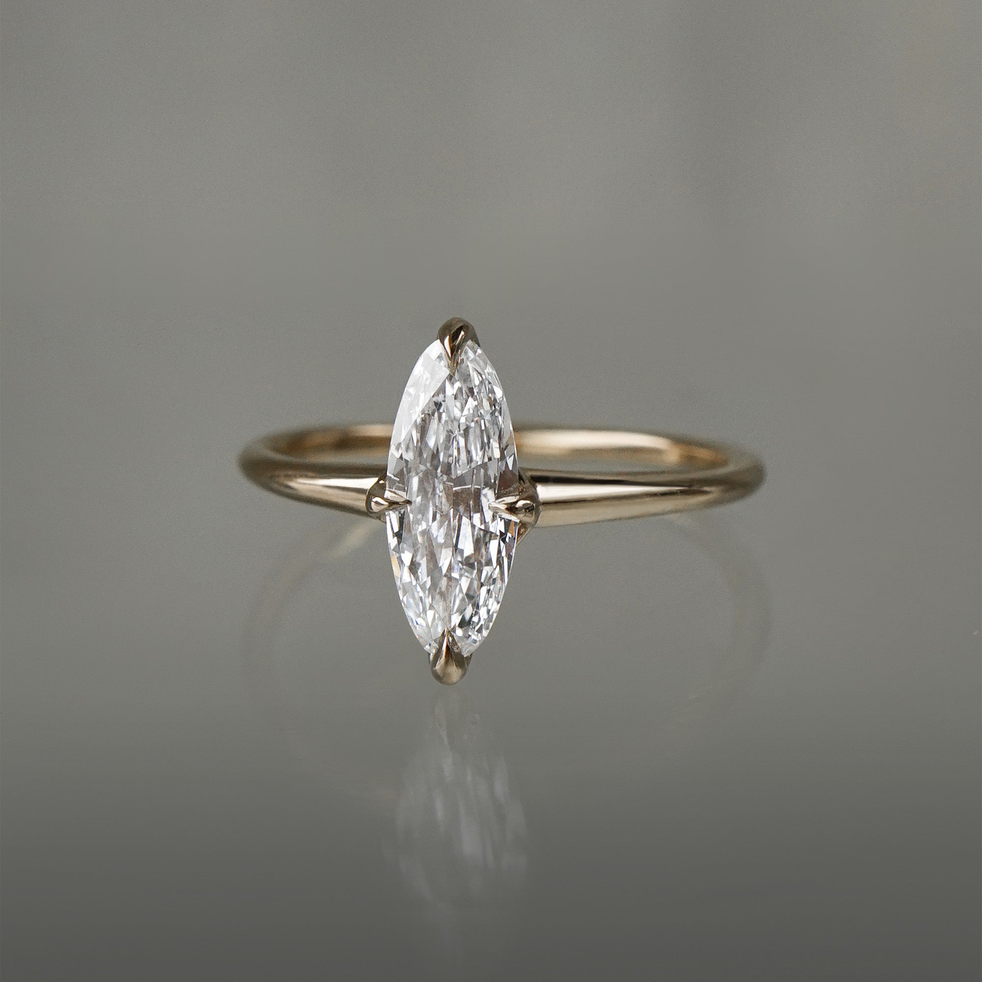 A one of a kind handcarved "Moondew" style engagement ring by Laurie Fleming Jewellery in solid 14k champagne gold. The ring features a unique moval shaped diamond (a long, marquise-oval mix) on a thoughtfully tapered plain gold band, held securely by four prongs at North, South, East, and West. The ring is situated on a medium grey background.