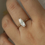 A hand wearing a one of a kind handcarved "Moondew" style engagement ring by Laurie Fleming Jewellery in solid 14k champagne gold. The ring features a unique moval shaped diamond (a long, marquise-oval mix) on a thoughtfully tapered plain gold band, held securely by four prongs at North, South, East, and West. The ring is held between an index finger and thumb, and rotated gently from side to side. The background is a light grey.