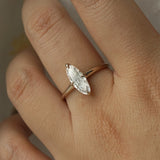 A hand wearing a one of a kind handcarved "Moondew" style engagement ring by Laurie Fleming Jewellery in solid 14k champagne gold. The ring features a unique moval shaped diamond (a long, marquise-oval mix) on a thoughtfully tapered plain gold band, held securely by four prongs at North, South, East, and West. The ring is held between an index finger and thumb, and rotated gently from side to side. The background is a light grey.
