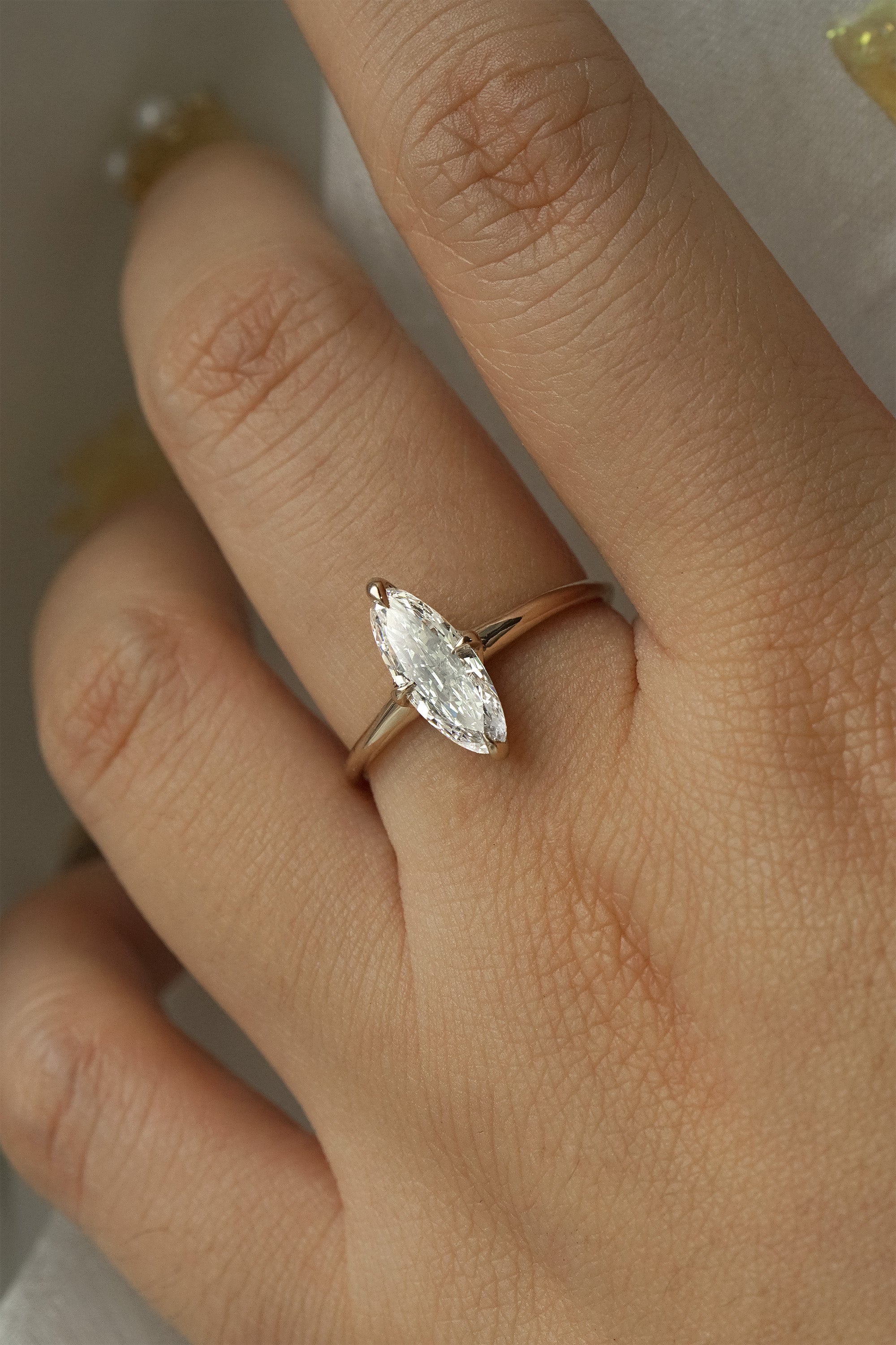 A hand wearing a one of a kind handcarved "Moondew" style engagement ring by Laurie Fleming Jewellery in solid 14k champagne gold. The ring features a unique moval shaped diamond (a long, marquise-oval mix) on a thoughtfully tapered plain gold band, held securely by four prongs at North, South, East, and West. The ring is held between an index finger and thumb, and rotated gently from side to side. The background is a light grey.