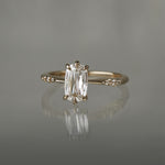 A delicate "Ilona" style one of a kind engagement ring by Laurie Fleming Jewellery in solid 14k champagne gold. It features a unique, elongated oval diamond with a "scissor cut", creating unique criss-crossed facets, held by six hand-shaped claw prongs. The band features small diamonds on the shoulders. The ring is situated on a grey background.