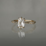 A delicate "Ilona" style one of a kind engagement ring by Laurie Fleming Jewellery in solid 14k champagne gold. It features a unique, elongated oval diamond with a "scissor cut", creating unique criss-crossed facets, held by six hand-shaped claw prongs. The band features small diamonds on the shoulders. The ring is situated on a grey background.