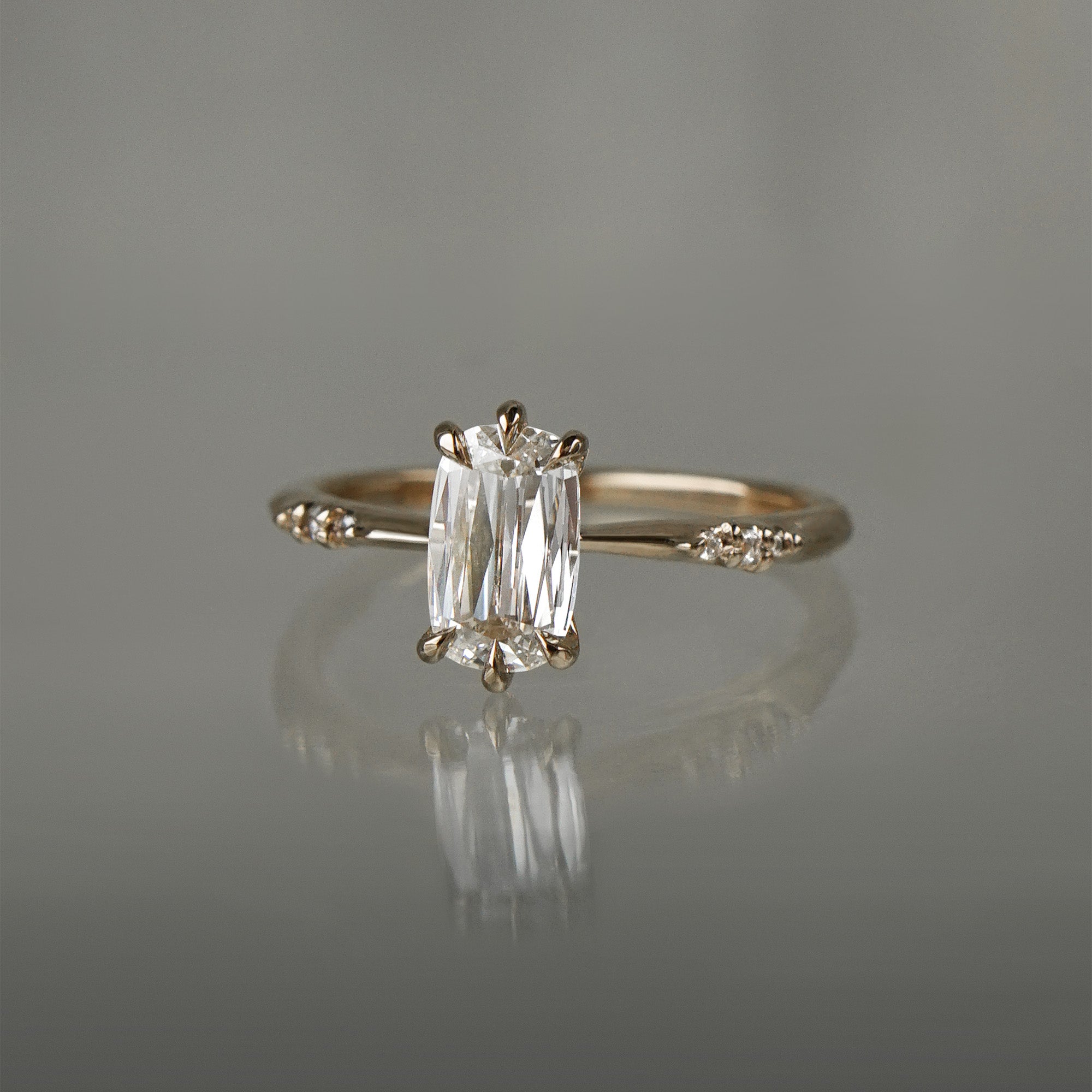 A delicate "Ilona" style one of a kind engagement ring by Laurie Fleming Jewellery in solid 14k champagne gold. It features a unique, elongated oval diamond with a "scissor cut", creating unique criss-crossed facets, held by six hand-shaped claw prongs. The band features small diamonds on the shoulders. The ring is situated on a grey background.