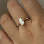 A delicate "Ilona" style one of a kind engagement ring by Laurie Fleming Jewellery in solid 14k champagne gold worn on a hand. It features a unique, elongated oval diamond with a "scissor cut", creating unique criss-crossed facets, held by six hand-shaped claw prongs. The band features small diamonds on the shoulders. The background is a light grey.