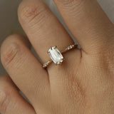 A delicate "Ilona" style one of a kind engagement ring by Laurie Fleming Jewellery in solid 14k champagne gold worn on a hand. It features a unique, elongated oval diamond with a "scissor cut", creating unique criss-crossed facets, held by six hand-shaped claw prongs. The band features small diamonds on the shoulders. The background is a light grey.