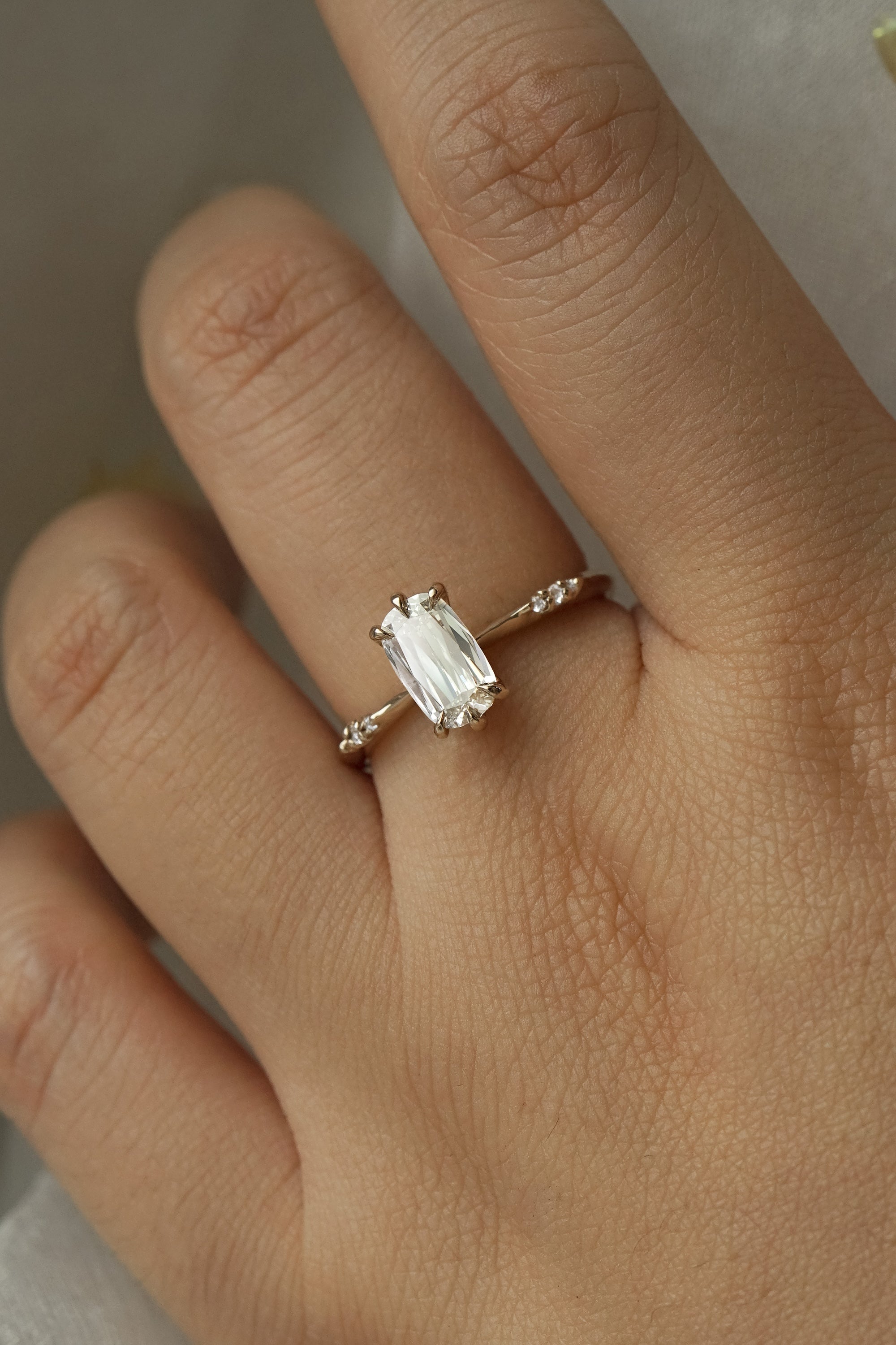 A delicate "Ilona" style one of a kind engagement ring by Laurie Fleming Jewellery in solid 14k champagne gold worn on a hand. It features a unique, elongated oval diamond with a "scissor cut", creating unique criss-crossed facets, held by six hand-shaped claw prongs. The band features small diamonds on the shoulders. The background is a light grey.