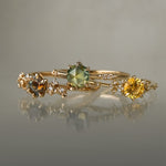 Three yellow gold rings on a grey background. On the left, a one of a kind "Asai Garden" ring with a round orange-blue sapphire centre and carved cherry blossoms on the band featuring diamonds. In the middle, an Ilona style ring with a round rose cut green sapphire on a delicate band with diamonds on the shoulders. On the right, a one of a kind Water Lily ring with a round bright yellow sapphire with clusters of diamonds on the band. All three rings are by Laurie Fleming Jewellery.