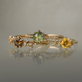 Three yellow gold rings on a grey background. On the left, a one of a kind "Asai Garden" ring with a round orange-blue sapphire centre and carved cherry blossoms on the band featuring diamonds. In the middle, an Ilona style ring with a round rose cut green sapphire on a delicate band with diamonds on the shoulders. On the right, a one of a kind Water Lily ring with a round bright yellow sapphire with clusters of diamonds on the band. All three rings are by Laurie Fleming Jewellery.