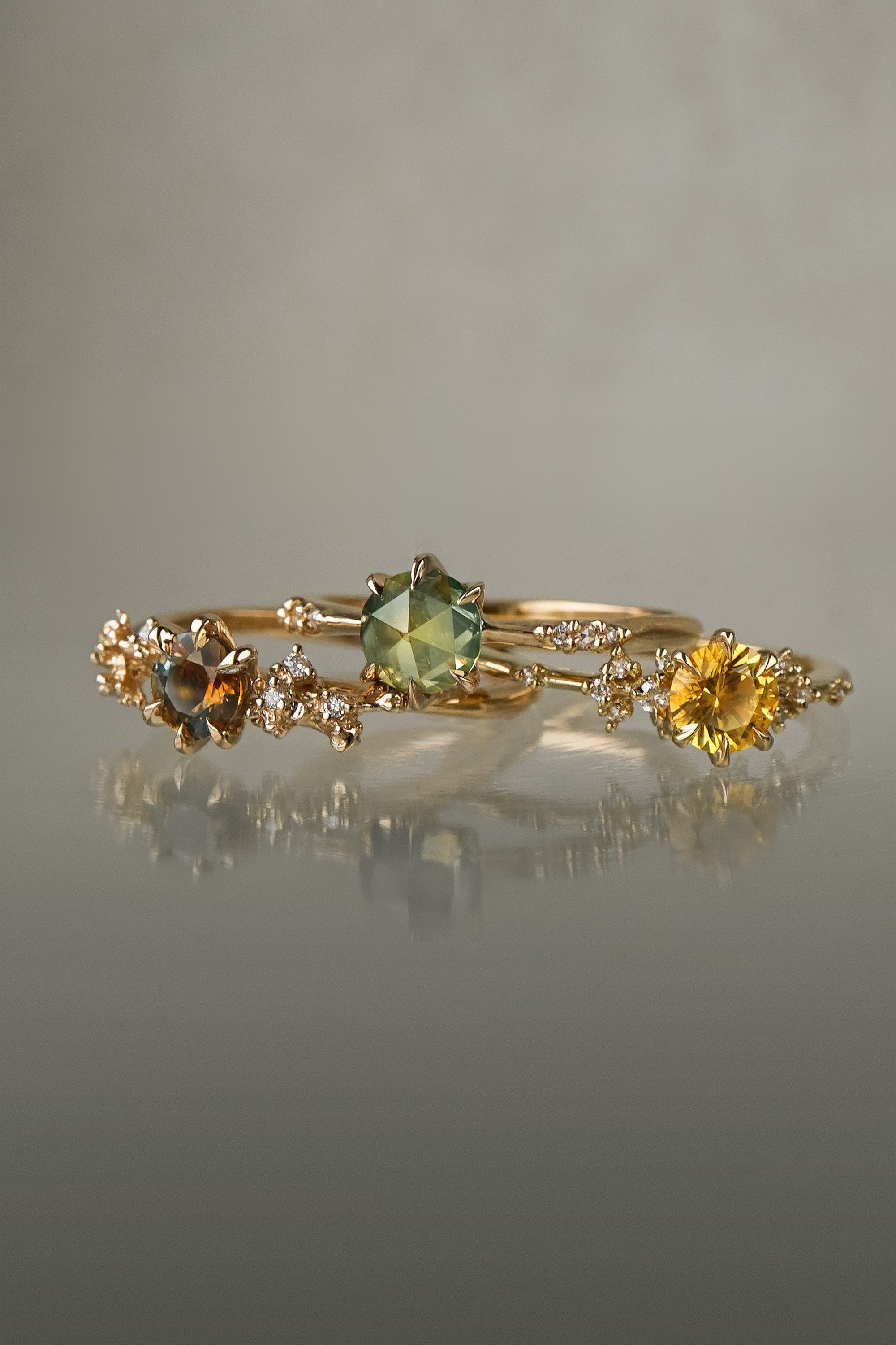 Three yellow gold rings on a grey background. On the left, a one of a kind "Asai Garden" ring with a round orange-blue sapphire centre and carved cherry blossoms on the band featuring diamonds. In the middle, an Ilona style ring with a round rose cut green sapphire on a delicate band with diamonds on the shoulders. On the right, a one of a kind Water Lily ring with a round bright yellow sapphire with clusters of diamonds on the band. All three rings are by Laurie Fleming Jewellery.