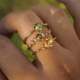 A hand wearing three yellow gold rings by Laurie Fleming Jewellery. The top ring is an Ilona style and features a round rose cut green sapphire on a delicate band with diamonds on the shoulders. In the middle, an Asrai Garden ring with a round orange-blue sapphire centre and carved cherry blossoms on the band featuring diamonds. At the base of the finger, a one of a kind Water Lily ring with a round bright yellow sapphire with clusters of diamonds on the band. The background looks like out of focus plants.