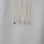 Four delicate yellow gold pendant necklaces by Laurie Fleming Jewellery draped over white fabric. From left to right: the blue-green teal Sapphire Moondew Necklace, the Rose Cut Mother of Pearl Moondew Necklace, the Rose Cut Diamond Moondew Necklace, and the Brilliant Cut Diamond Moondew Necklace. - Laurie Fleming Jewellery